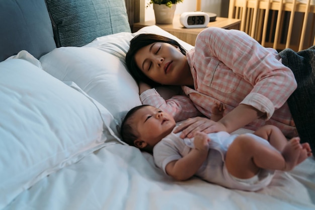 tired asian new mom wearing pajamas is falling asleep on the bed while comforting her waking baby at night during bedtime in the bedroom.