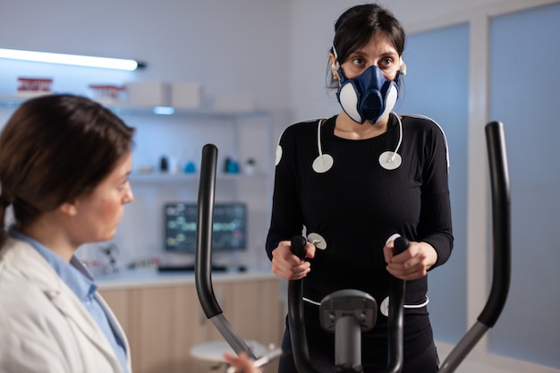 Tired athlete woman training on cross trainer increasing, rhythm of exercises wearing mask and medical electrodes monitoring muscle endurance and cardiac rate in science sport laboratory.