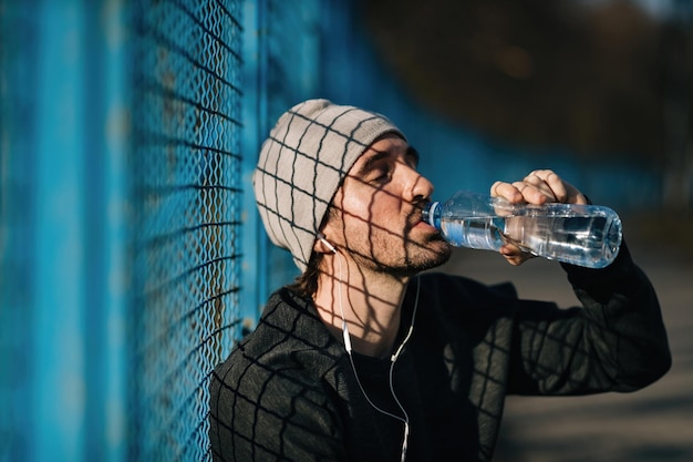 Усталый спортивный мужчина пьет воду с закрытыми глазами, отдыхая от занятий спортом на открытом воздухе.
