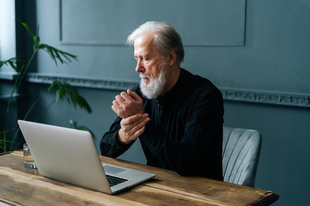 Tired bearded mature adult male working typing on laptop\
experiences pain in wrist, carpal tunnel syndrome. overworked\
gray-haired older man working on pc and suffering from pain in\
hand.