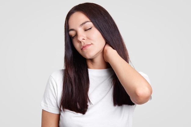 Tired beautiful brunette female keeps hand on neck, feels pain, tired after working in office, dressed in casual white t shirt, isolated on studio wall. Fatigue woman feels sick. onworking concept