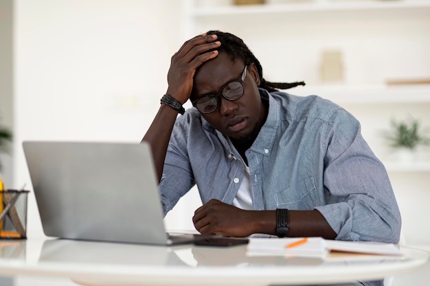 Tired black man sitting at desk with laptop and touching\
head