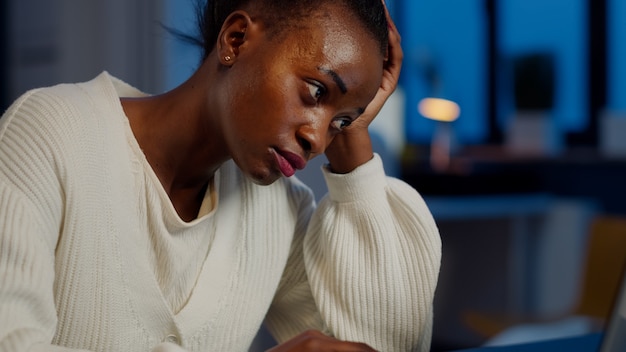 Tired black woman almost napping on chair working on laptop in start up company office late at night. Busy employee using modern technology doing overtime to finish project, respecting deadline