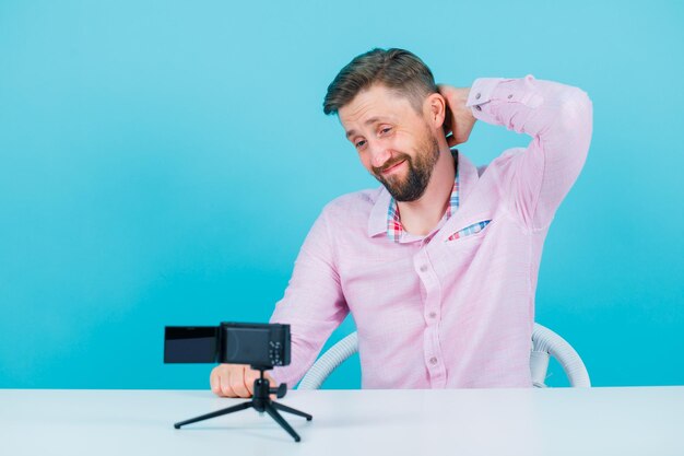 Tired blogger man is holding ahnd behind head by sitting in front of his mini camera on blue background