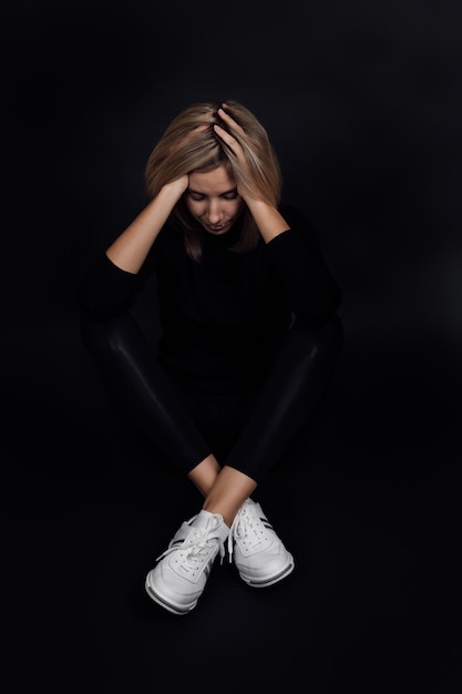 Tired blonde woman with head down and eyes closed hands\
touching hair sitting on floor on black background physical and\
mental abuse relative aggression