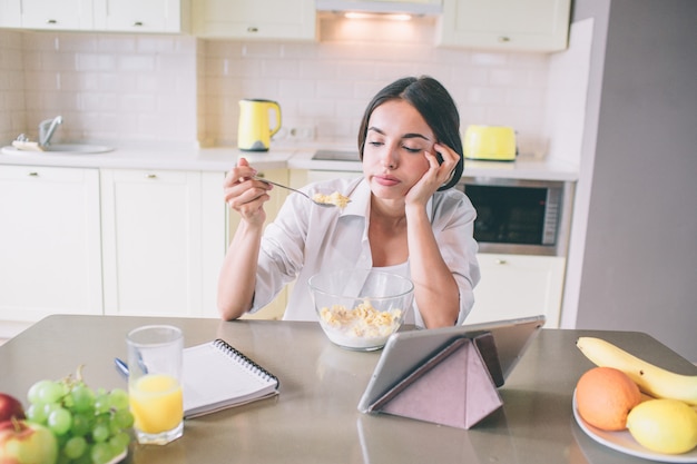 Tired and bored girl is sitting at table and looking at spoon with milk and corn flakes.