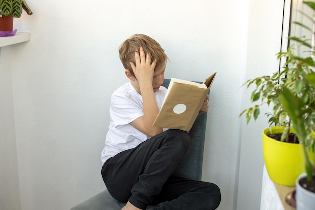 Tired boy sits on a chair with a book and holds his head with his hand