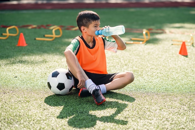 Tired boy in soccer uniform drinks with water after intensive training