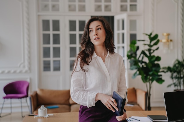 Tired brunette young woman in white shirt and violet pants sits on table looks aside with tired face