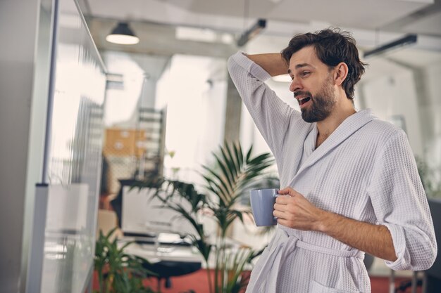 Tired businessman in white bathrobe looking at whiteboard and yawning while drinking hot morning beverage in office