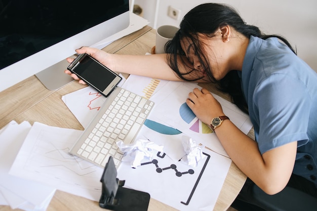 Tired businesswoman sleeping on the desk while holding smartphone in front of the computer screen