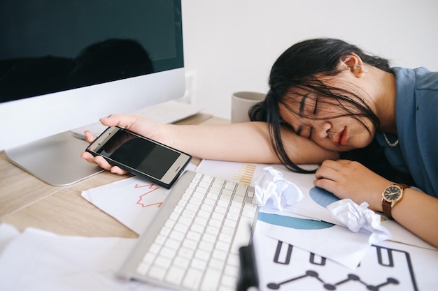 Tired businesswoman sleeping on the desk while holding smartphone in front of the computer screen