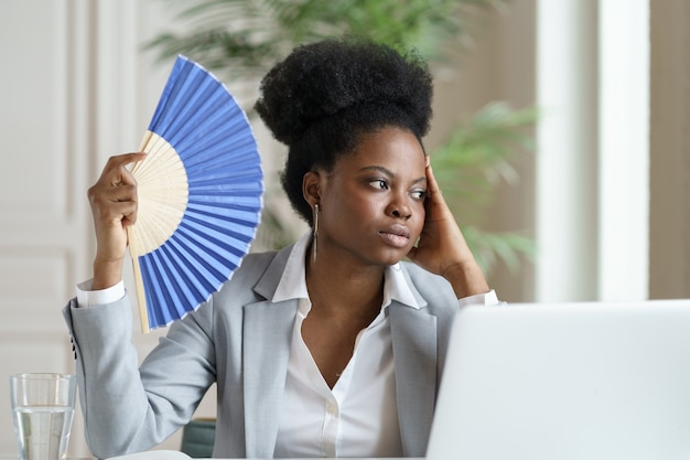 Tired businesswoman waving paper fan exhausted of heat overheated at workplace in office or at home