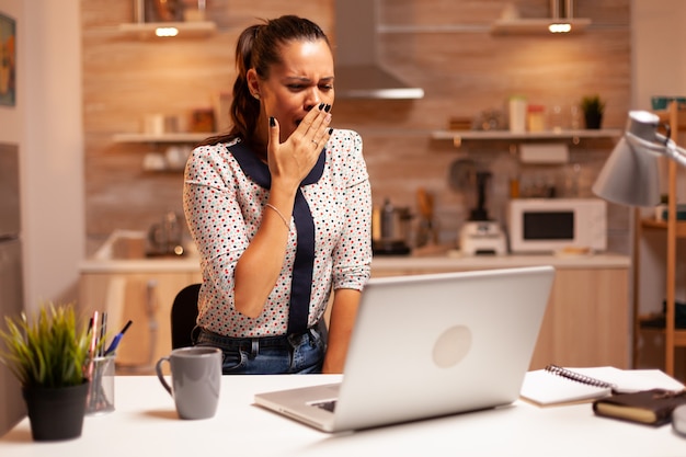 Tired businesswoman yawning while working on a deadline using laptop in home kitchen. Employee using modern technology at midnight doing overtime for job, business, busy, career, network, lifestyle.