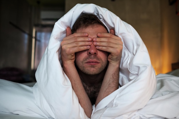 The tired caucasian man lay on the bed and cover his face with arms