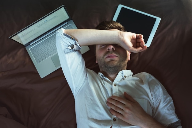 Tired caucasian young man in white shirt sleeping and keeping one hand above head lying on bed near laptop