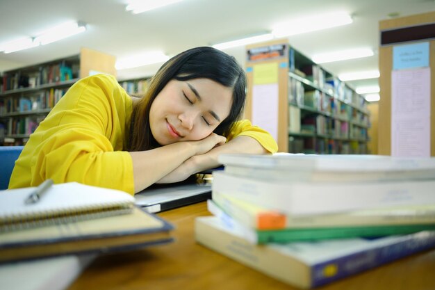 背景にぼやけた本棚を持つ図書館の本の山で寝ている疲れた大学生