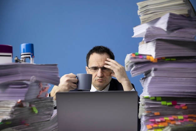 Tired concerned businessman holding cup of coffee at office desk with huge pile of documents