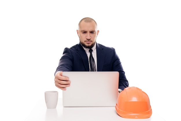 A tired constructor businessman in an orange helmet looks at the laptop screen and studies the construction project. Sits at the table and drinks coffee, nervous