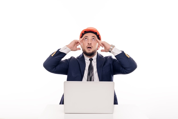 A tired constructor businessman in an orange helmet looks at the laptop screen and studies the construction project. Sits at the table and drinks coffee, nervous