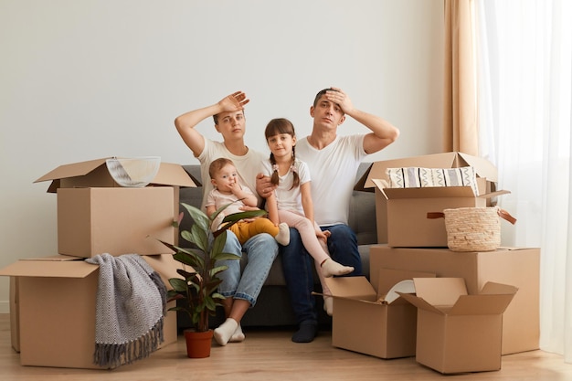 Tired couple moving to a new home with their children, sitting on cough with exhausted expression, keeping hands on foreheads, unpacking personal piles, relocation.