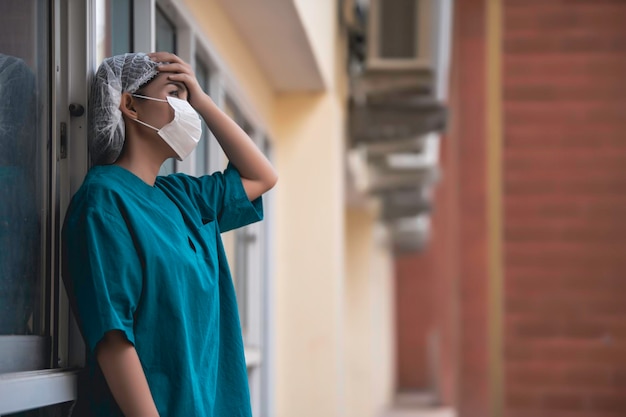 Tired depressed female asian scrub nurse wears face mask blue uniform sits on hospital floorYoung woman doctor stressed from hard work