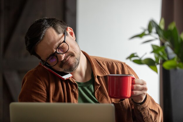 Tired developer working with laptop, drink coffee and calling by mobile phone at home