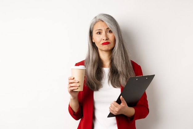 Tired and disappointed asian businesswoman with grey hair, drinking coffee in paper cup and looking gloomy at camera, standing over white background