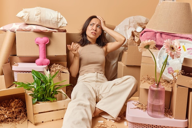 Tired displeased asian woman dressed in t shirt and trousers\
lies around cardboard boxes relocates to new apartment surrounded\
by cardboard boxes full of personal belongings has much work to\
do