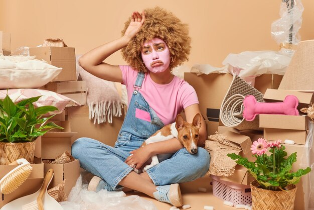 Tired displeased curly haired young woman applies beauty mask\
on face feels exhausted after relocation and packing personal\
belongings in cardboard boxes poses with pet on floor in lotus\
pose