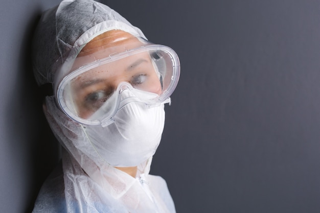 A tired doctor in medical goggles mask and protective overalls looks sadly at the camera