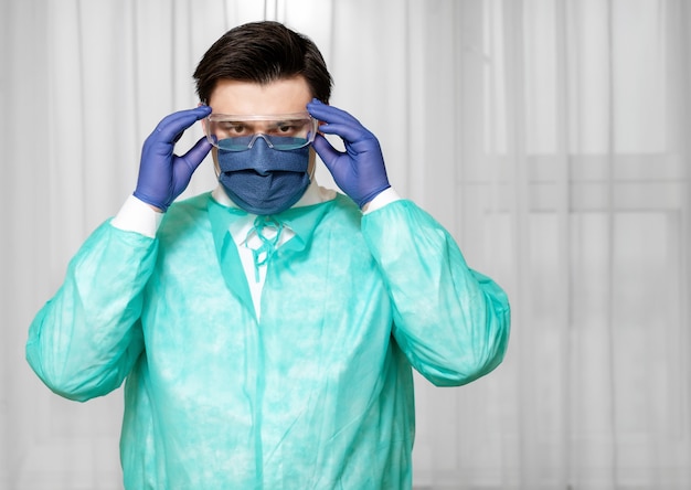 Tired doctor in protective equipment at home puts on protective medical mask with glasses, coronavirus epidemic