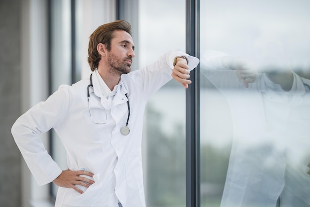 A tired doctor standing near a window and looking on the watch in an empty hospital hallway during the Covid-19 pandemic.