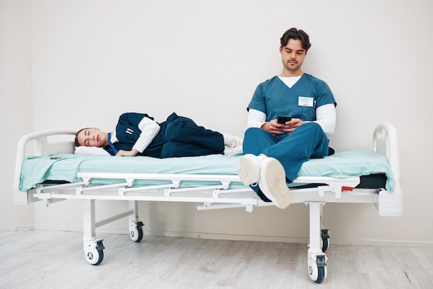 Photo tired doctors woman and man in hallway with phone texting and relax on bed at hospital job medic team partnership or friends with burnout smartphone and fatigue with healthcare clinic and rest