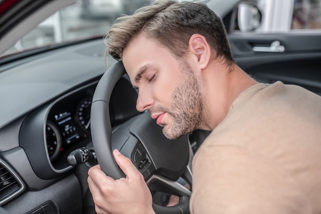 疲れたドライバー。車のハンドルに頭を乗せた軽いTシャツを着た居眠りしている男は日中疲れています