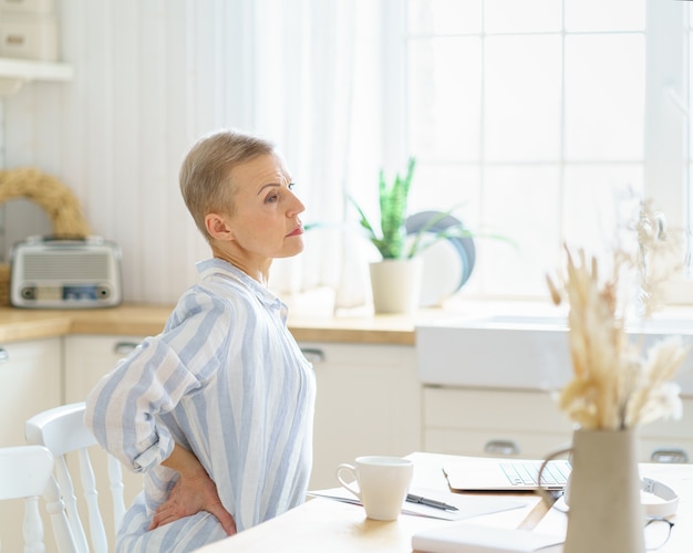 Tired exhausted middle aged business woman massaging her back while sitting in kitchen and working