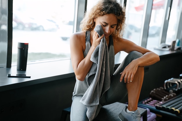 Tired female athlete after an active body workout rests wiping sweat with a towel in the gym concept...