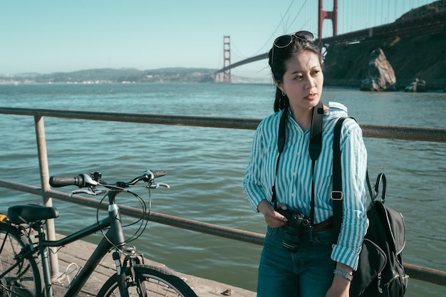 Tired female backpacker resting relying on iron metal handrail by the coastline near the blue ocean sea. red golden gate bridge in background under sunny day. girl lens man leisure relaxing with bike