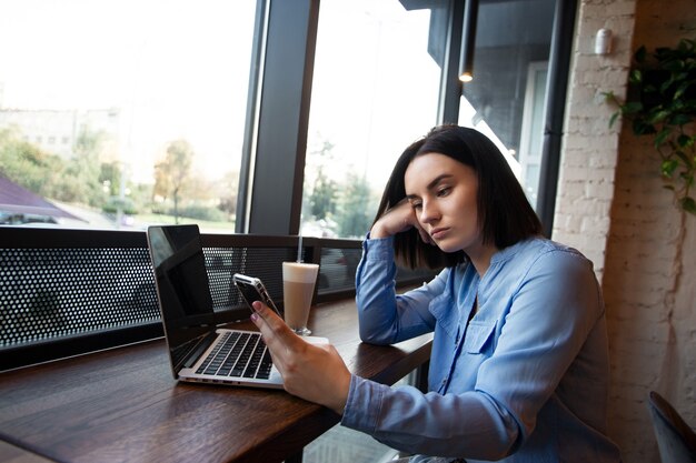 カフェで働く疲れた女性フリーランサー。集中問題の概念。スマートフォンを見て、電話を待っている動揺した若い女性。居心地の良いコーヒーショップの雰囲気。背景にカフェやレストラン。