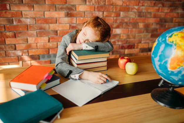 Tired female pupil asleep on the stack of textbooks at the table with opened notebook.