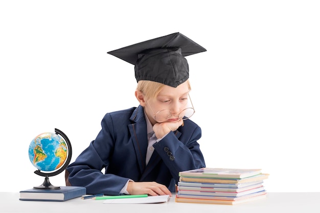 Stanco ragazzo di prima elementare nel cappello degli studenti fa i compiti scolaro annoiato isolato su sfondo bianco concetto di istruzione