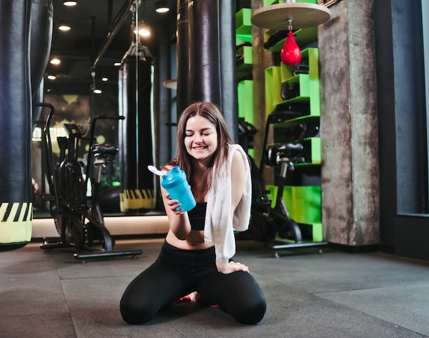 Tired fit girl in sportswear sits on the floor and holds bottle of water with towel