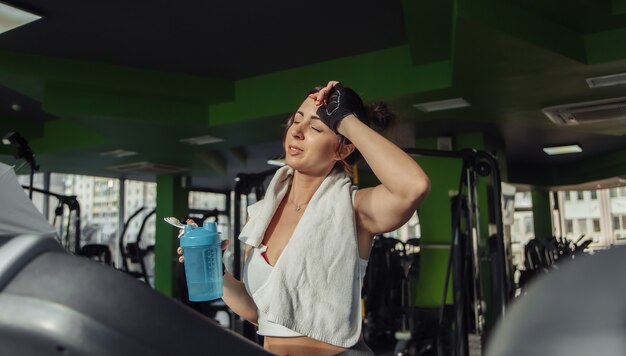 Tired fit woman with a towel on her shoulders holding water bottle on a treadmill. Weight Loss Concept, Aerobic Workout