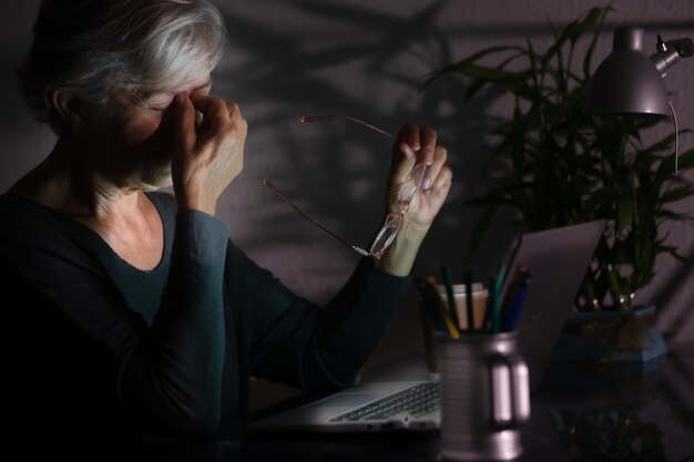 夜遅くにラップトップを使用して作業している疲れて欲求不満の年配の女性。