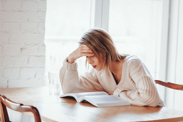 Ragazza stanca in maglione bianco con il libro alla cucina