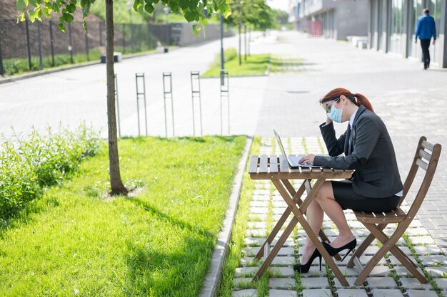 眼鏡をかけた疲れた女の子が屋外でコンピューターでリモート作業している マスクをしたビジネスウーマンがコロナウイルスの流行中にサマーテラスに座りながらラップトップでタイプしている 社会的距離