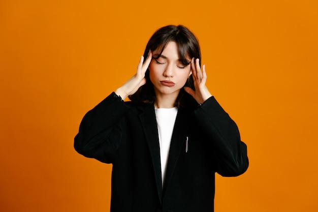 Tired grabbed head young beautiful female wearing black jacket isolated on orange background