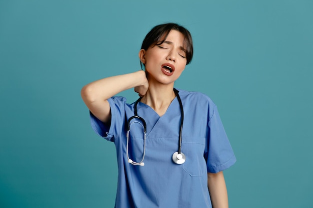 Усталая схватившаяся за шею молодая женщина-врач в униформе, стетоскоп, изолированный на синем фоне