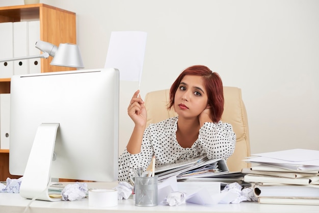 Tired hopeless pretty young businesswoman leaning on stack of documents and lifting hand with white ...