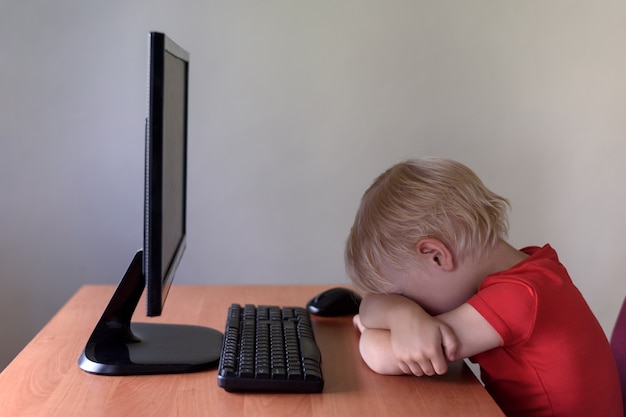 Tired little blond boy sleeping on a table under the monitor